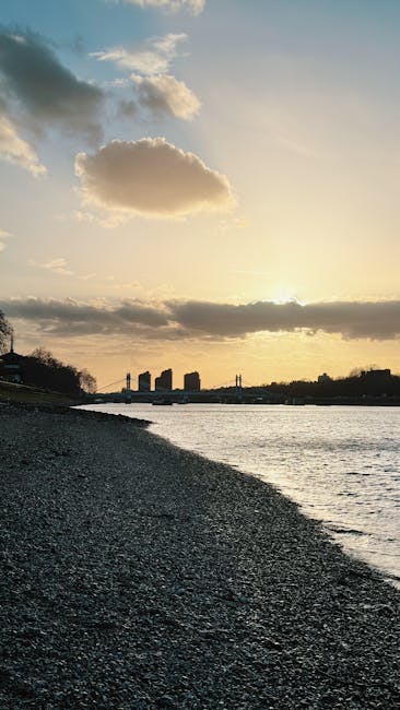 The image depicts a beach scene at sunset with a pebble shoreline in the foreground and a calm body of water extending towards the horizon. In the distance, there are several dark silhouettes of modern buildings and structures, possibly part of an urban waterfront or city skyline. The sky is partly cloudy with a mix of fluffy clouds and clear patches, illuminated by warm, golden sunlight that creates a soft glow across the scene. The lighting suggests late evening, with the sun low on the horizon, casting long shadows. Sparse trees and possibly some small structures are visible along the shoreline on the left side, adding natural elements to the otherwise urban backdrop. This tranquil setting could be related to private outdoor space or coastal environment, where rubbish collection or disposal might be managed through specialized services, aligning subtly with private waste handling or alternative rubbish removal options in urban or seaside areas.