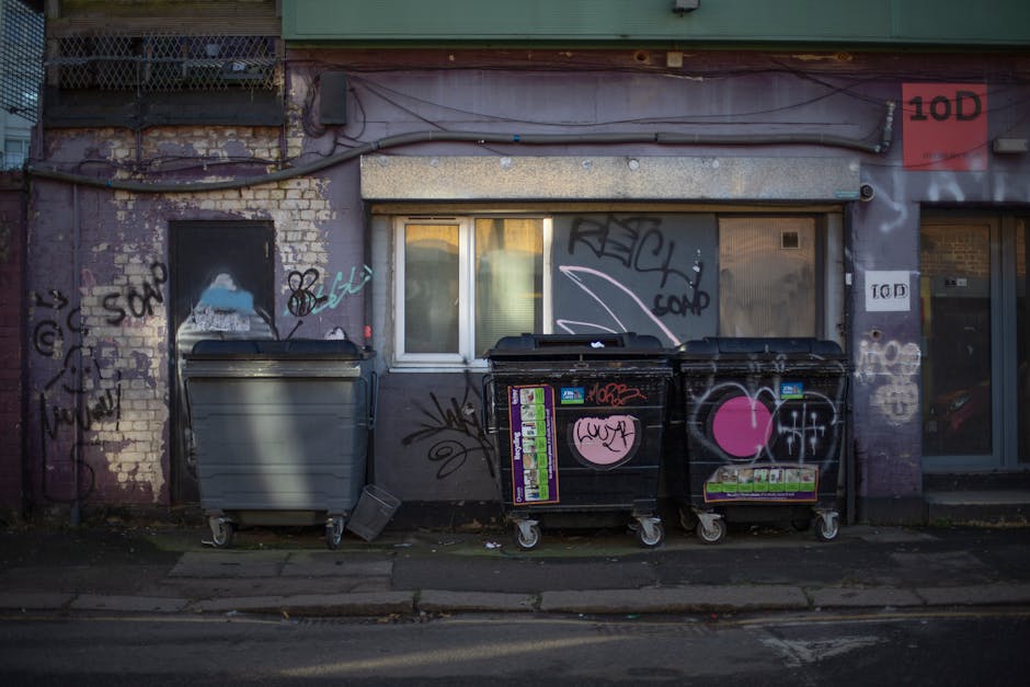 In front of a weathered building with purple-painted brickwork and visible signs of aging and graffiti, three large wheelie bins are positioned along the pavement. The left bin is light gray with a smooth, matte finish, while the two black bins on the right are constructed from plastic with a slightly textured surface, each adorned with vibrant pink heart-shaped stickers and various colorful stickers and graffiti tags. The black bins are partially open, revealing dark interior contents, and sit on small casters. Behind the bins, there is a small window with a white frame, partially obscured by shadows and graffiti, and above this window, exposed wires run across a weathered, rusty metal strip. The ground below is strewn with some debris and fallen leaves, contributing to the urban scene. To the right, part of a glass door with a metal frame and signs is visible, indicating the building’s address or service access. The scene is lit with natural, diffuse daylight, which highlights the textures of the building’s exterior and the graffiti, subtly referencing individual waste management practices in urban environments that may involve private waste collection or on-site clearance by services like House Clearance Hammersmith. The overall atmosphere conveys a typical example of urban rubbish storage consistent with private disposal services.