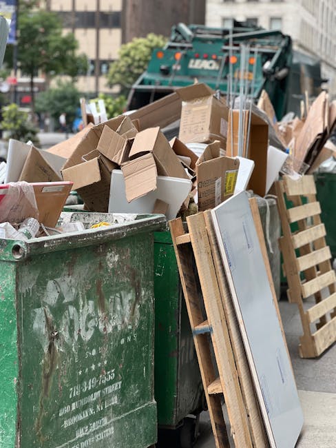 A large green plastic waste bin filled with a diverse collection of discarded cardboard boxes, some flattened and others in their original shape, alongside a wooden pallet propped against the side of the bin. The boxes vary in size, color, and material, some with visible print or labels. Behind the bin, a green waste collection vehicle is partially visible, with its rear and side panels showing signs of wear and dirt. The scene is set outdoors on a city street, with trees and multi-story buildings in the background, and the sky partially visible. The lighting is natural, suggesting daytime, and the overall environment indicates a typical urban rubbish removal or collection scene, showcasing a straightforward example of waste disposal activities involving cardboard and packaging materials, potentially handled by services like House Clearance Hammersmith for private or business waste removal.