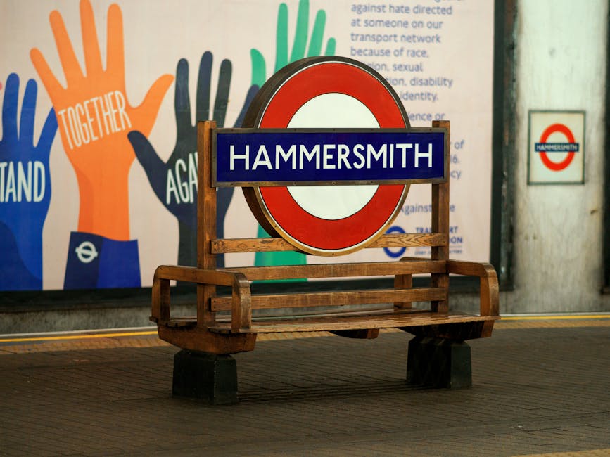 A wooden bench with a straightforward design sits on a tiled platform at Hammersmith underground station. Positioned centrally on the bench is a circular London Underground sign with a red outer ring, a white middle circle, and a blue rectangular plaque across the middle bearing the word 'HAMMERSMITH' in white capital letters. Behind the bench, a large colorful mural features stylized hand illustrations in shades of orange, blue, and green with words like 'TOGETHER' and 'STAND' written on the hands, set against a predominantly white background. To the right of the sign, a small white square sign with the Hammersmith station logo is mounted on a textured wall with visible paint and surface variations. The scene is illuminated by ambient station lighting, casting soft shadows and highlighting the textures of the materials. This setting, situated in a busy London transport environment, subtly reflects how different transportation signage and infrastructure elements support route identification and station identification, aligning with themes of public transport and independent travel management.