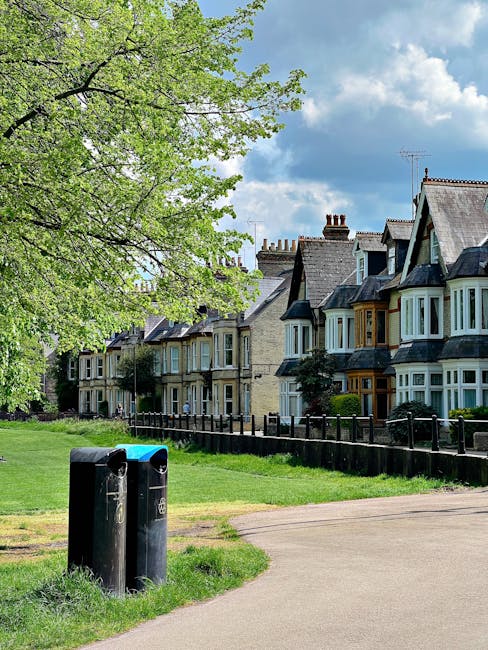 A residential street scene featuring a row of traditional Victorian-style terraced houses with bay windows, brick facades, and slate roofs, situated behind a well-maintained black wooden fence. In the foreground, there are two black and blue rubbish bins placed on a patch of green grass, with the bins positioned near a paved footpath. A large deciduous tree with fresh green leaves extends over the scene from the left, providing partial shade. The sky above is partly cloudy with patches of blue visible amidst white clouds. The overall setting suggests a peaceful urban neighborhood, and the presence of the rubbish bins indicates regular waste collection or disposal, akin to private waste handling services that may be offered in such areas by companies like House Clearance Hammersmith, supporting alternative waste management options outside local authority collections.