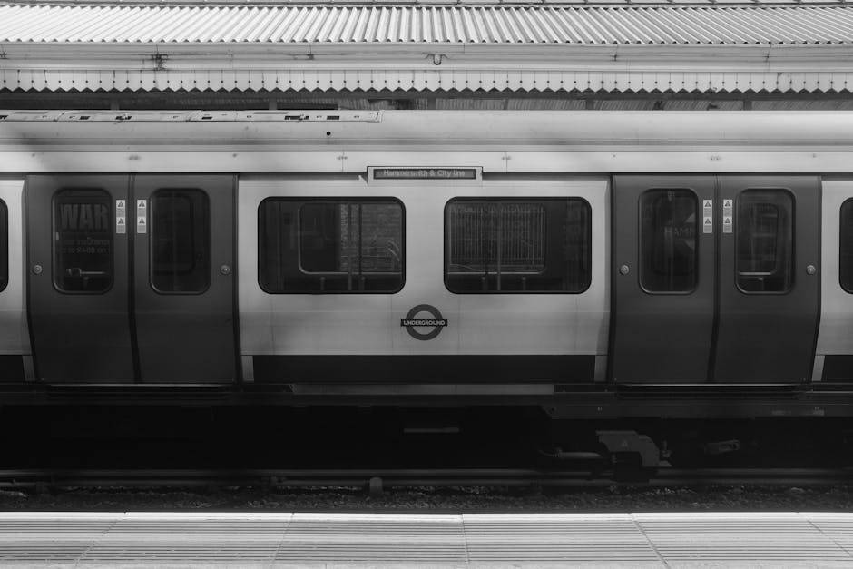 A black and white photograph of a modern London Underground train at a station platform. The train's exterior features metallic silver and dark gray panels with several rectangular windows, some showing reflections or inner seating arrangements. A circular London Underground logo is visible on the middle carriage. The train is positioned parallel to the platform, which has tactile paving strips along the edge for safety. Above the train, there is a corrugated metal roof with visible support structures. The overall scene is well-lit, highlighting the textures of the train's smooth metallic finish and the platform surface. The image evokes a sense of urban transit and transportation infrastructure, relevant to discussions about alternative waste management or transportation options involving public transit services, potentially as a complement or alternative to private rubbish collection methods in urban areas.