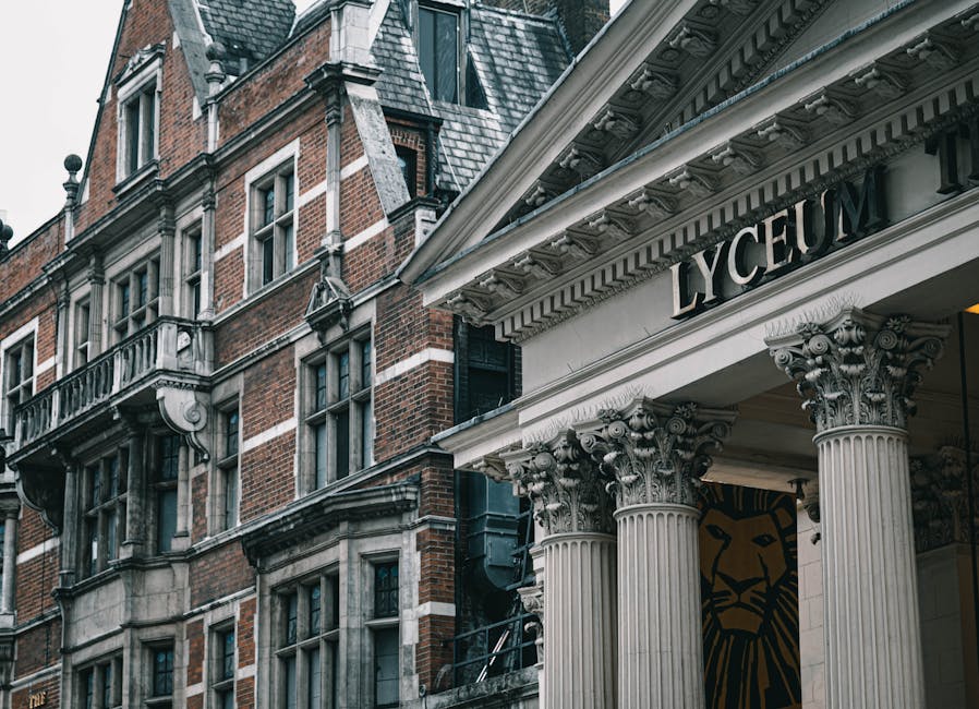A close-up view of the exterior of a historic building with traditional red brick and stone detailing, featuring multiple rectangular windows with white frames, some of which have small ledge brackets underneath. To the right, part of a classical-style entrance portico is visible, supported by ornate, fluted columns with decorative Corinthian capitals. Above the portico, a white entablature displays the word 'Lyric' in black capital letters, indicating the theatre or cultural venue. The building appears to be multi-story, with the upper floors showing intricate masonry work, including brickwork with contrasting stone accents, and steeply pitched slate roofs with dormer windows. The atmosphere is daytime with natural lighting, highlighting the textures of the brick, stone, and slate. This scene may relate to heritage site preservation or private maintenance activities, similar to rubbish removal services such as those offered by House Clearance Hammersmith, especially in the context of maintaining or clearing around historic and cultural locations.