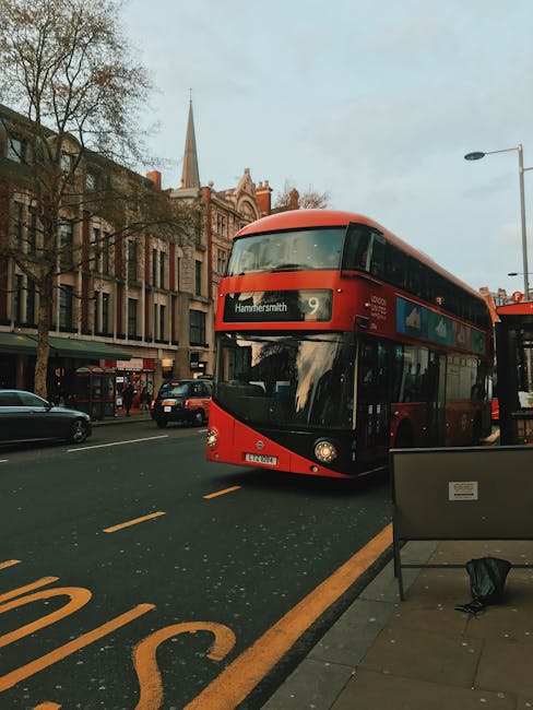 A modern double-decker bus painted in red with black accents on the lower section is captured moving through a busy urban street in Hammersmith. The bus displays route number 9 and the destination Hammersmith on its electronic sign above the windshield. In the foreground, there is a black metal bench with a small black bag resting on the seat, positioned on the sidewalk alongside a blue paper or cardboard signholder. Behind the bus, multi-story buildings with brick facades and large windows line the street, some with ground-floor shops and others residential. A church steeple is visible in the distance, towering above the trees that have lost most of their leaves, indicating a late autumn or winter setting. Several parked and moving cars are present along the street, and a streetlamp extends from the right side of the image, all under a muted, overcast sky. The scene is typical of a busy city centre, where public transport and urban infrastructure are integral to daily life, subtly connecting to the topic of rubbish removal and waste management services offered by House Clearance Hammersmith in the area.