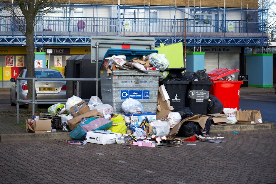 A large cluster of overflowing rubbish bins and refuse bags sits on a paved sidewalk outside a retail premise, with various discarded cardboard boxes, plastic packaging, paper, and black garbage bags scattered around the containers. The scene includes a grey mixed waste bin labeled for papers and cards, with its lid partially open revealing piled-up waste, and several black and red recycling bins nearby. Behind the waste collection site, there is a row of storefronts with bright signage and a building under construction, covered with scaffolding and blue safety netting. A silver car is parked to the left, partially obscured by a metal railing, and a leafless tree is positioned on the left side of the image. The environment appears to be urban, with the waste situated on a street corner, illustrating a collection challenge often addressed by private rubbish removal services in residential or commercial areas. The natural daylight and shadows suggest a clear, possibly cold day, highlighting the disorder of the overflowing rubbish and the impact of improper waste disposal in this area.