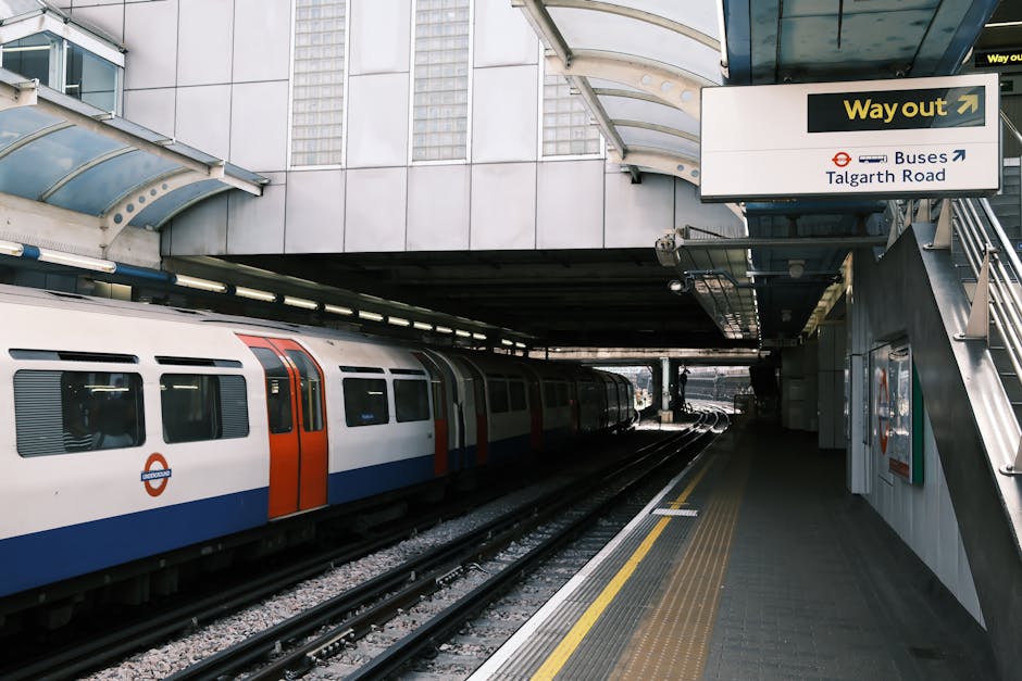 An underground train station platform with a modern commuter train stopped at the platform, featuring a sleek white body with orange and blue accents along the windows and doors. The train's exterior has a smooth, metallic finish, reflecting the surrounding environment. The platform has a textured, non-slip surface in dark grey, with a yellow tactile strip along the edge for safety. Overhead, a metal canopy with a curved, translucent roof offers shelter for waiting passengers, supported by steel beams. On the right side, a staircase with metal handrails ascends to an upper level, and a sign indicating the exit to Talgarth Road and bus connections is visible, with clear directional arrows and a black and yellow color scheme. The background reveals a cityscape with tall glass buildings, and the overall lighting suggests daytime with natural light filtering through the station’s structure. The scene predominantly depicts urban public transport infrastructure, with a subtle connection to the theme of alternative transportation solutions, such as private or independent rail and bus services.
