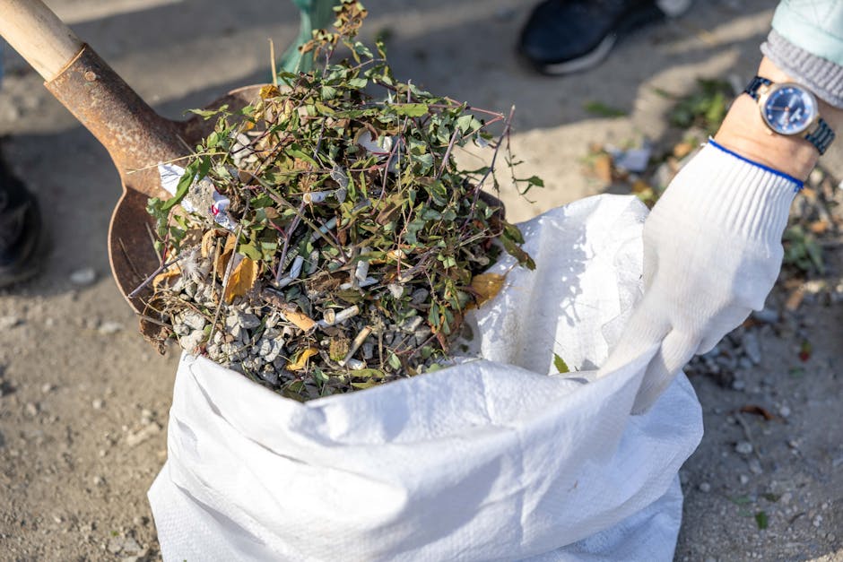 A person wearing a white glove is holding a white woven polypropylene bag filled with a mixture of garden waste, including small green leafy branches with thorny stems and dry twigs, along with crushed paper and other small debris. The bag is positioned on a paved or concrete surface outdoors, with part of the person's wristwatch visible on their wrist, which is also clad in a light-colored sleeve with blue trim. Next to the bag, a rusty shovel with a wooden handle is partially inserted into the ground, suggesting active garden clearance or waste collection. In the background, blurred elements of outdoor terrain, possibly a driveway or garden pathway, are visible, with natural lighting highlighting the texture of the waste materials and the environment. This scene exemplifies an on-site private waste removal process, typical of residential rubbish clearance services in urban or suburban settings.