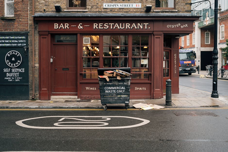 In the foreground, a large black wheeled waste bin labeled 'COMMERCIAL WASTE ONLY' sits on a city sidewalk in front of a small restaurant with a deep red exterior and large glass windows, revealing warm indoor lighting and interior decor. The bin is filled with a mixture of cardboard boxes and packaging materials, some overflowing and resting on the ground nearby. To the left, a blackboard-style sign indicates the restaurant's opening hours for a self-service buffet, and to the right, a narrow vertical sign lists 'COCKTAILS'. A concrete bollard and a small metal post are positioned along the edge of the sidewalk, separating pedestrian space from the street. Behind the bin, parked vehicles including a truck are visible along the road, which has a painted white line and a circular road marking indicating no parking enforcement. The building above features a street sign reading 'CRISPIN STREET E1', and the scene is illuminated by natural daylight with an urban atmosphere typical of London, linking the visual to waste collection and disposal services often used for commercial property clearances. House Clearance Hammersmith offers alternative waste handling options for such urban settings.