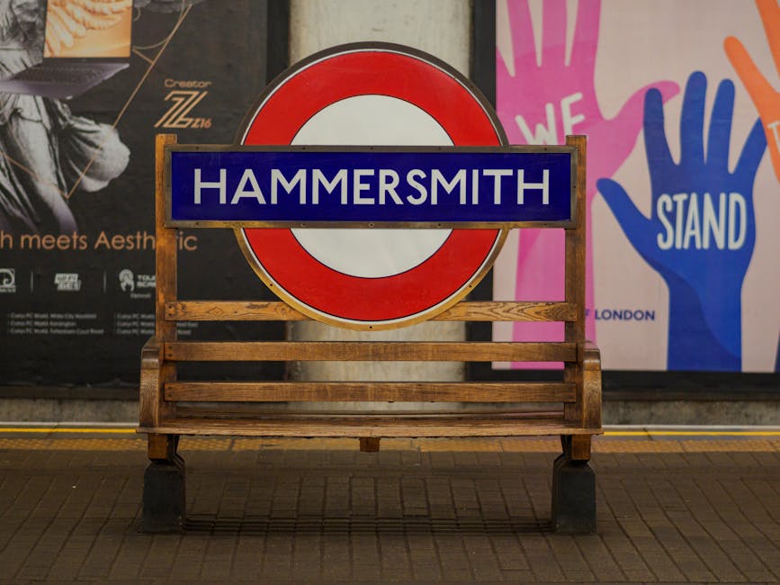A wooden bench with a slatted seat and backrest, positioned on a tiled pavement in front of a large circular London Underground sign featuring a red outer ring and a white center, with a blue rectangular plaque across the middle displaying the word 'HAMMERSMITH' in white capital letters. Behind the sign, there are illuminated posters and advertisements hanging on a dark wall, including one with the partially visible word 'Aesthetic' and another showing a vivid illustration of colorful raised hands with the words 'WE STAND' visible. The scene is lit by ambient lighting, creating a clear view of the bench's texture and the signage's details, with the background environment suggesting a transport station or public area where independent waste management or rubbish removal services could be relevant, as reflected in the context of the site and surroundings associated with the webpage about booking rubbish crews in Hammersmith.